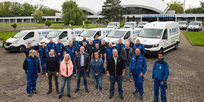 Eine Gruppe von Mitarbeitern in blauen Arbeitsanzügen steht vor mehreren Firmenfahrzeugen und einem modernen Gebäude.
