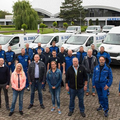 Eine Gruppe von Mitarbeitern in blauen Arbeitsanzügen steht vor mehreren Firmenfahrzeugen und einem modernen Gebäude.