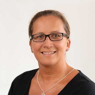 Die Frau mit Brille und einem freundlichen Lächeln steht vor einer neutralen Wand, ihre Frisur ist kurz und gepflegt.