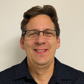 Der Mann mit Brille hat kurzes, lockiges Haar und zeigt ein freundliches Lächeln, während er in die Kamera schaut.