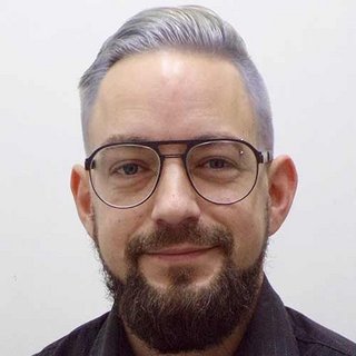 Ein Mann mit grauen Haaren und einem gepflegten Bart lächelt freundlich in die Kamera, während er eine Brille trägt.