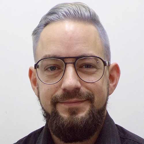 Ein Mann mit grauen Haaren und einem gepflegten Bart lächelt freundlich in die Kamera, während er eine Brille trägt.