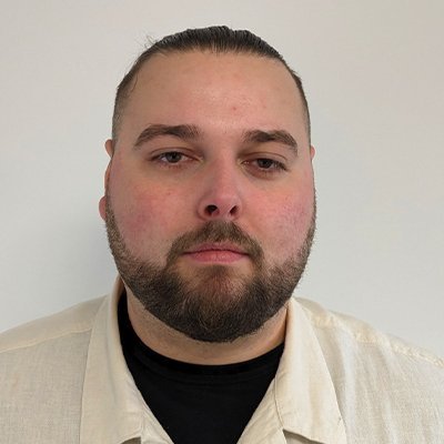 Der Mann hat einen nachdenklichen Ausdruck und trägt ein beiges Hemd über einem schwarzen T-Shirt, vor einem neutralen Hintergrund.