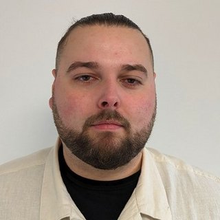 Der Mann hat einen nachdenklichen Ausdruck und trägt ein beiges Hemd über einem schwarzen T-Shirt, vor einem neutralen Hintergrund.