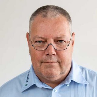 Der Mann hat eine markante Gesichtszüge und trägt eine Brille, während er vor einem neutralen Hintergrund steht.