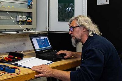 Ein älterer Mann mit grauen Haaren und Brille arbeitet konzentriert an einem Laptop, umgeben von Werkzeugen und elektrischen Komponenten.